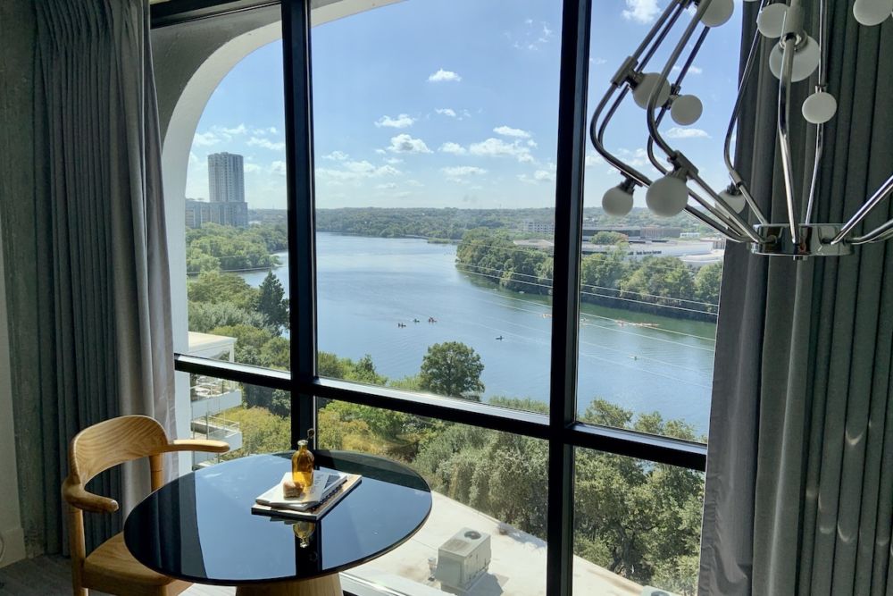 Looking out the window from a room at the boutique hotel The Line. View is of the Colorado River in Austin, and there is a steel lamp with glass baubles hanging in the right corner. A small round table and wooden chair are in front of the window.