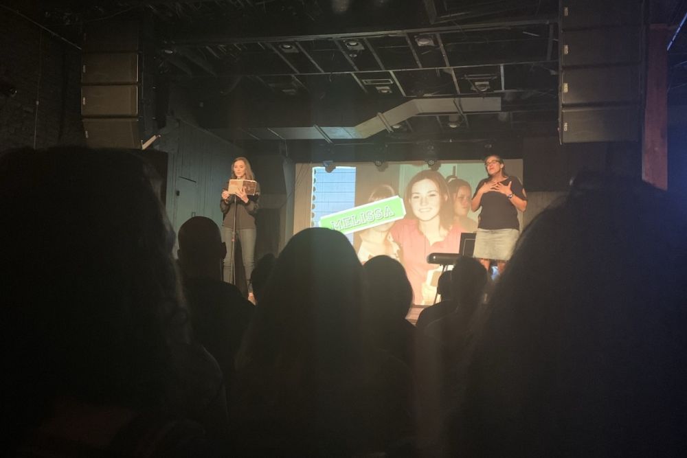 A woman in her 20s or 30s dressed in a black long-sleeved shirt and jeans reads her diary from the stage. A female ASL interpreter stands to her right. Behind them is a large screen with a picture of the diarist as a teen.
