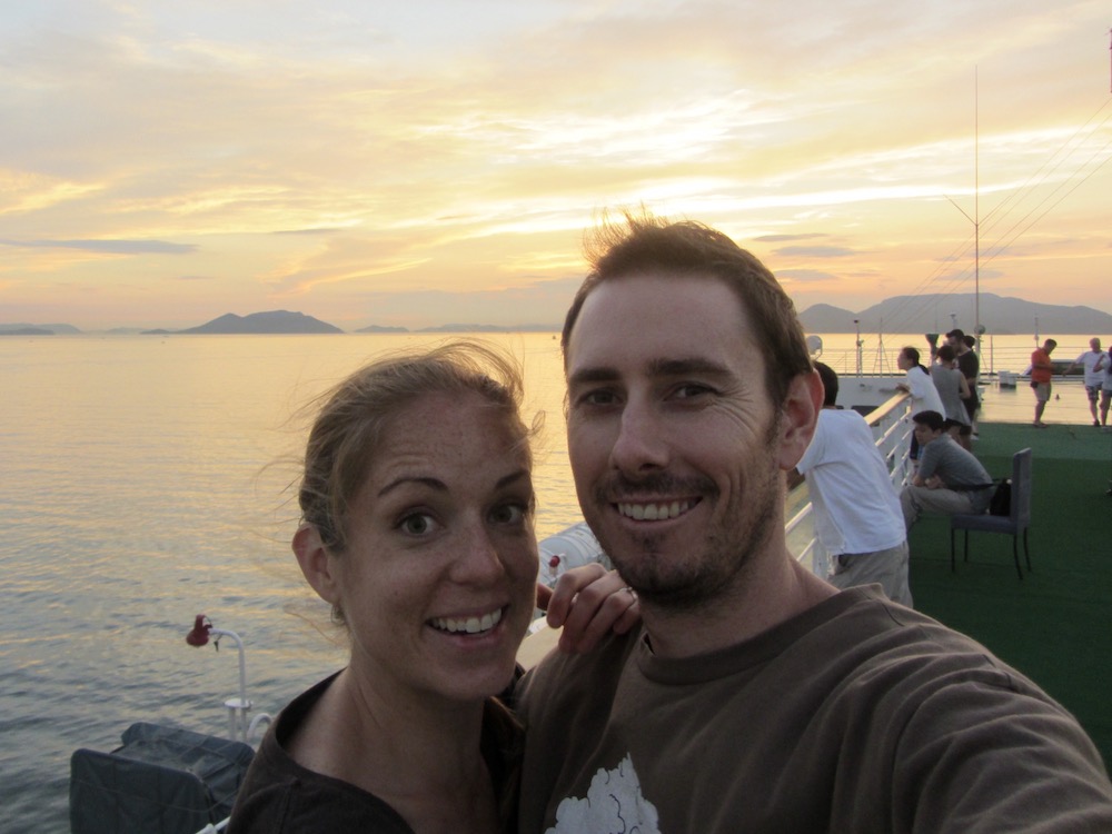 Man and woman take a selfie from the roof of a ferry as the sun sets behind them. Her face is surprised, he is smiling and bearded. 