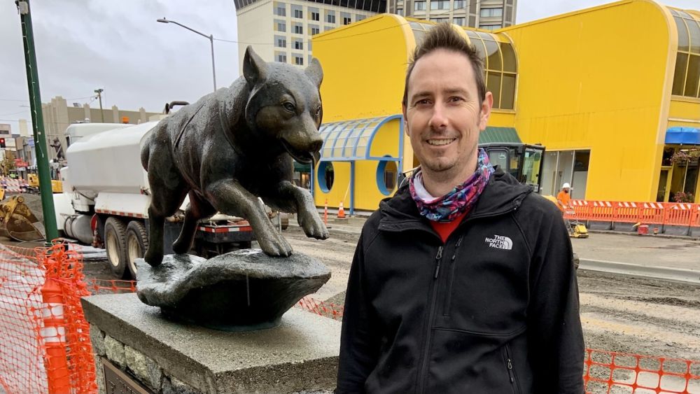 Man smiling in front of sled dog statue in downtown Anchorage