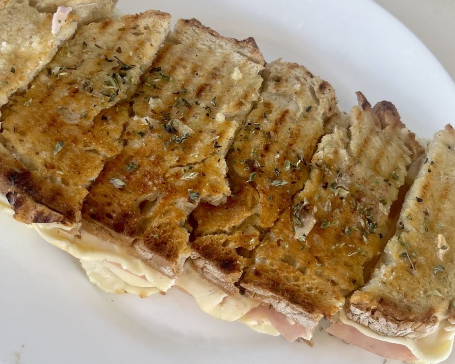 A large toasted ham and cheese sandwich with fresh oregano sprinkled on top is cut into six rectangular slices on a white plate.