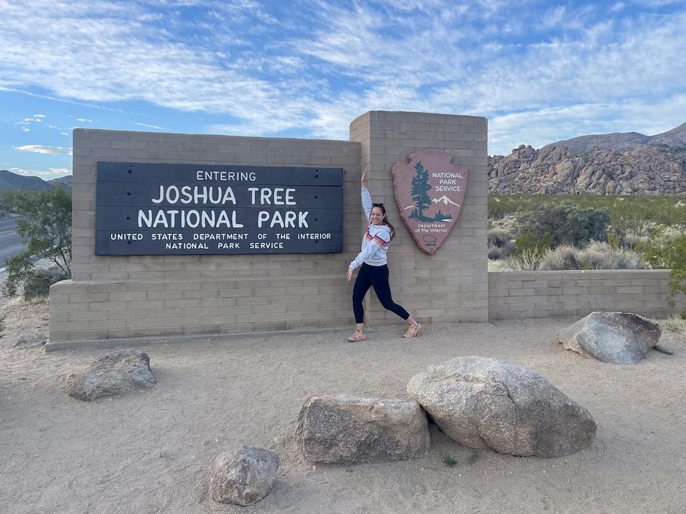 Lauren wears a grey hoodie with rainbow stripes and leggings as she proudly stands next to the "Entering Joshua Tree National Park" sign.