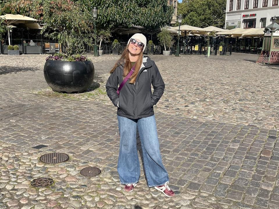 Lauren wears very wide-legged jeans, a grey rain jacket, a cream beanie, and merlot colored tennis shoes. She stands on cobbled streets in a city square in Malmo, Sweden. 