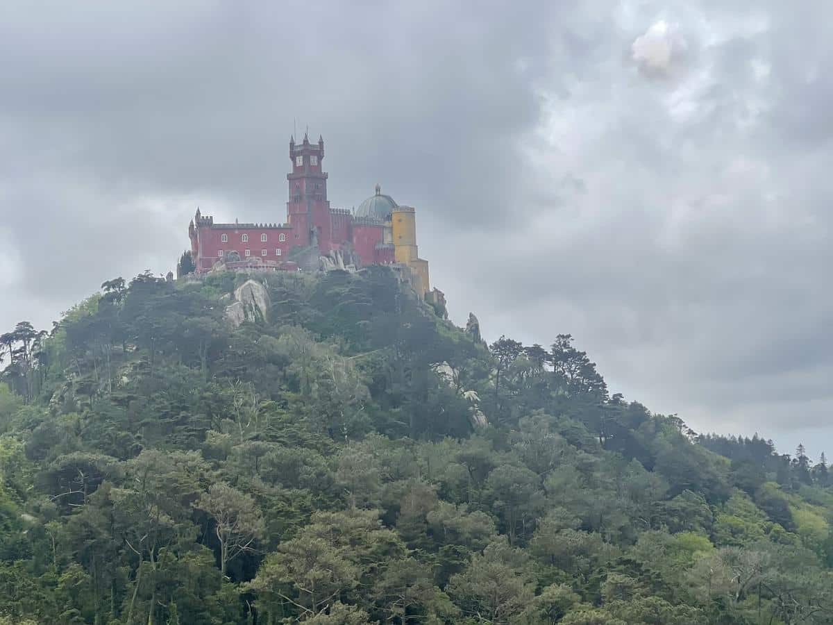 The red and yellow castle of Pena palace, the most popular attraction for a day trip to Sintra, viewed from below. A slight gauzy mist covers the castle as it sits on a green wooded hill.