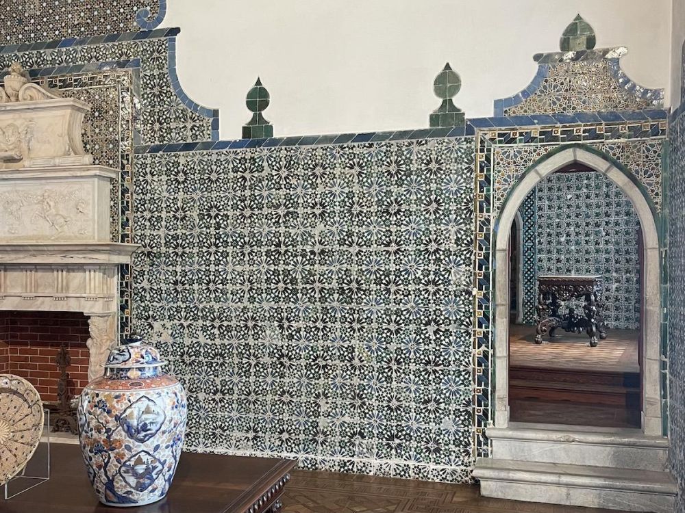 intricately detailed tilework next to an arched doorway inside the National Palace of Sintra. We can see part of a fireplace and a wooden table and decorative vase in the foreground.