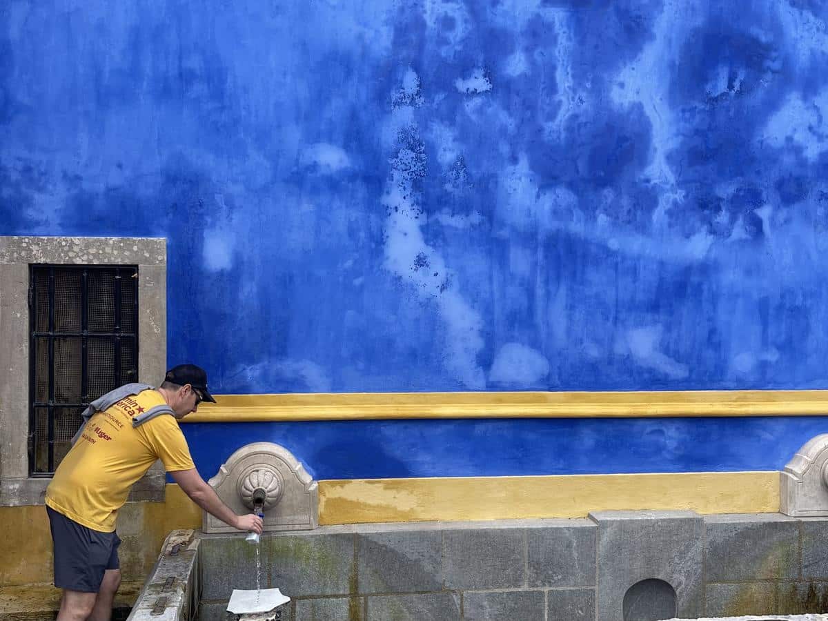A man in a yellow t-shirt and black hat fills up a water bottle at a decorative public water fountain that has a large bright blue wall.