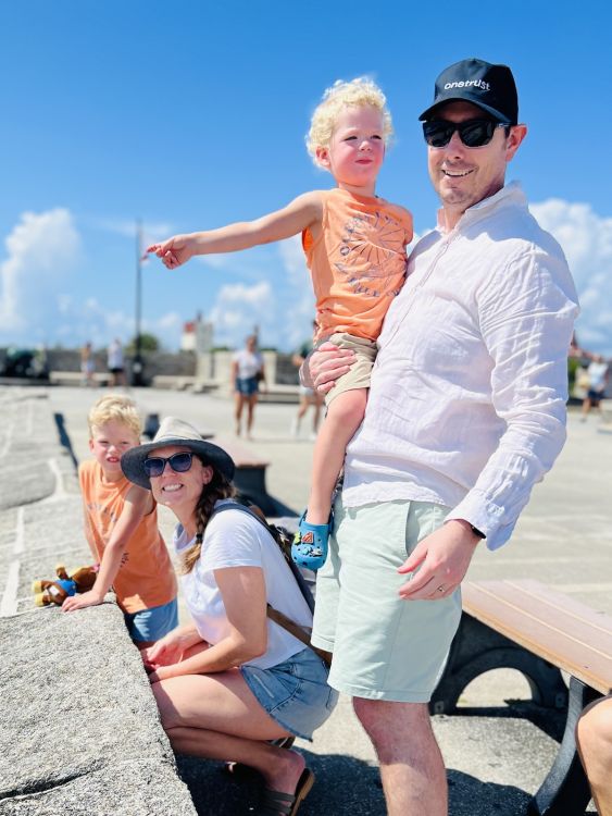 An man and a women dressed in white tops and shorts hold two toddlers in matching orange t-shirts. They're on top of the fort in St. Augustine, Florida. 