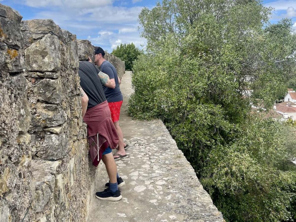 Two middle-aged white men lean between the turrets of the wall in Óbidos. They stand on a narrow ledge with no barriers or guardrails. A large tree is behind them.