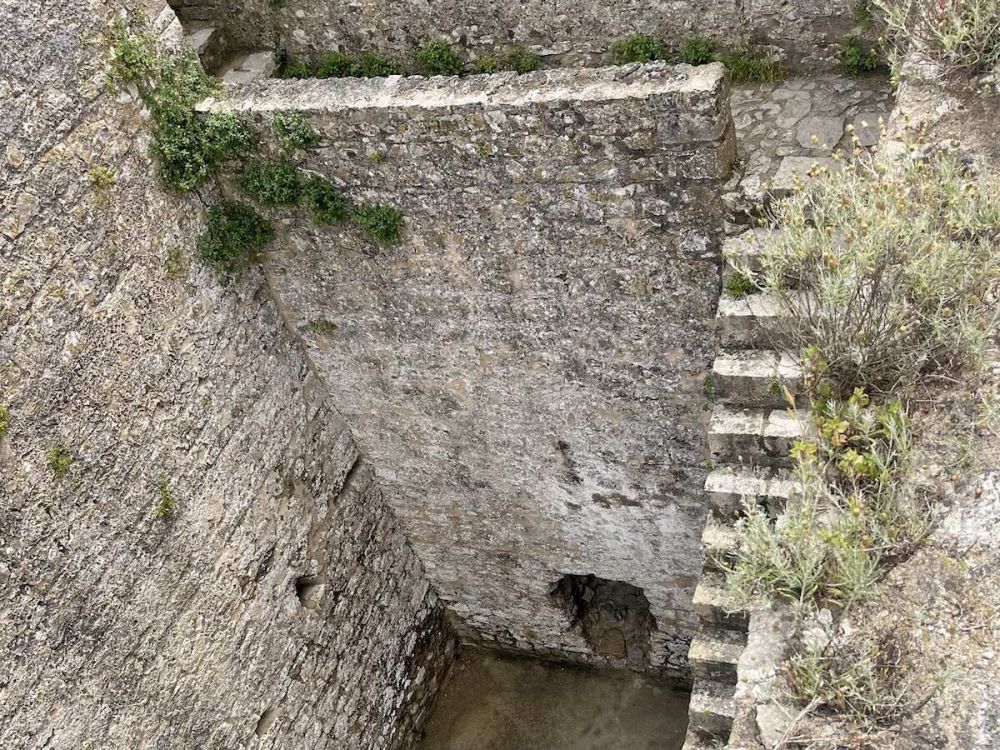 A steep stone staircase runs along the inner edges of a stone tower. There are no handrails and shrubs line the edge of the photo.