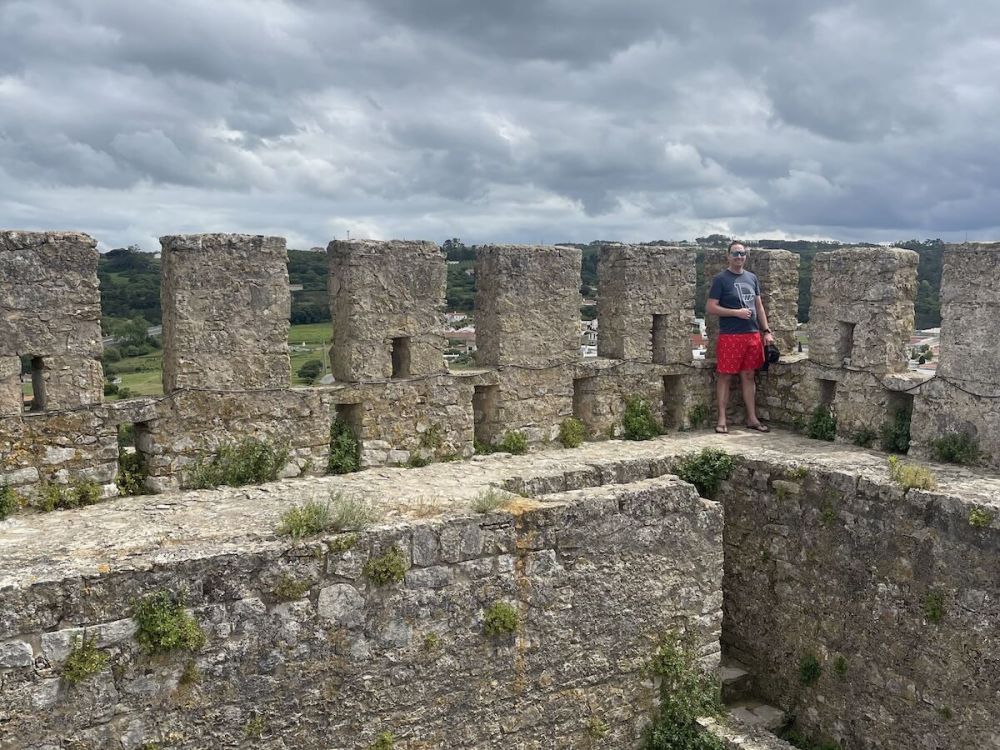 A white man in sunglasses and red swim trunks grins as he stands in the corner of a tower on a ledge. He has a hat in his hand and wears flip flops.