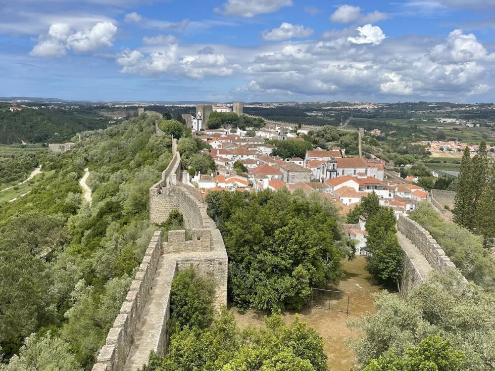 View from the city walls of Óbidos, Portugal. The town is filled with white walled buildings and orange rooftops. Rolling green hills surround it and the sky is blue with fluffy white clouds.
