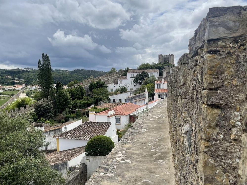 View from the narrow ledge on the wall of Óbidos, Portugal. A stone tower is visible in the background and several white buildings are inside the city walls. A storm gathers in the distance.