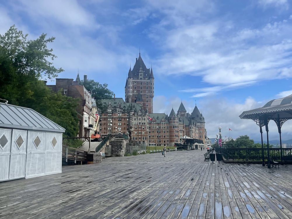 The chateau Fairmont looms over a boardwalk. The sky is blue but small puddles on the wood indicate recent rain. 