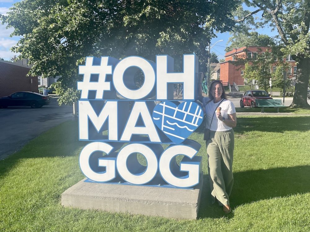 A white woman in a white t-shirt and green linen pants holds an ice craem cone and smiles next to a six foot sign that says OH MA GOG.