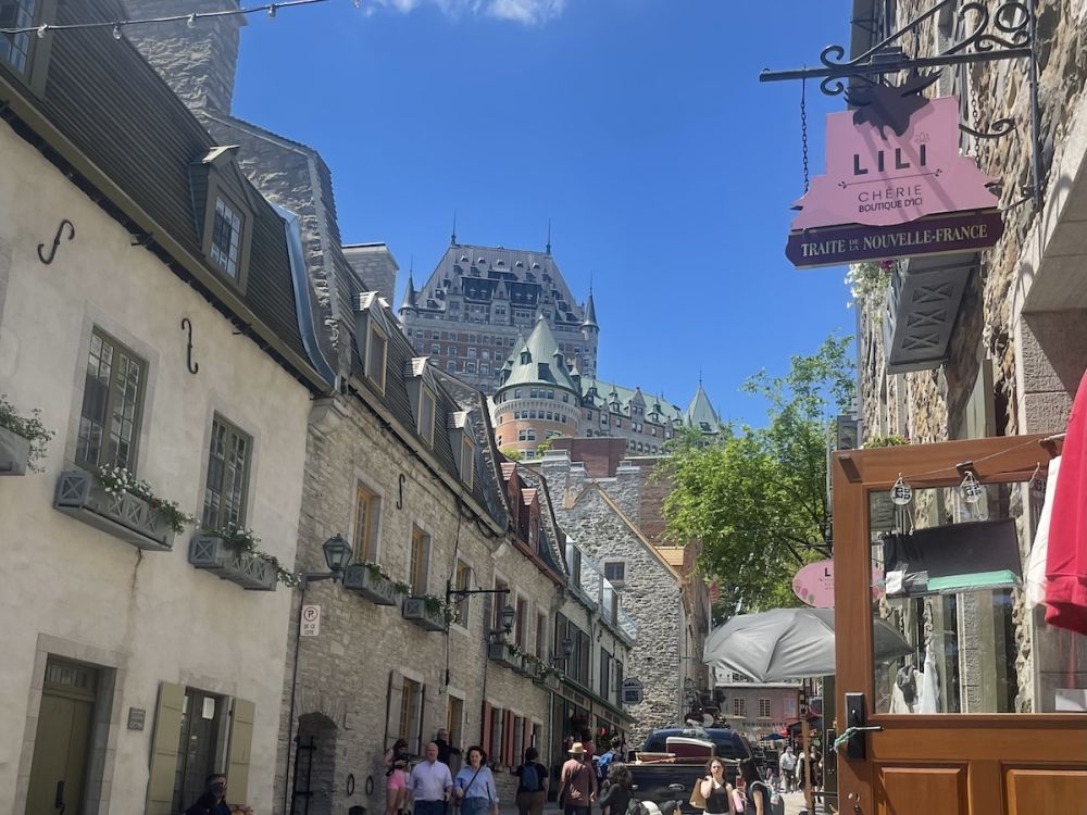 A quaint street in old town Quebec City with shops along the edge, tourists in the road, and the chateau-style hotel Fairmont in the background above it all. 