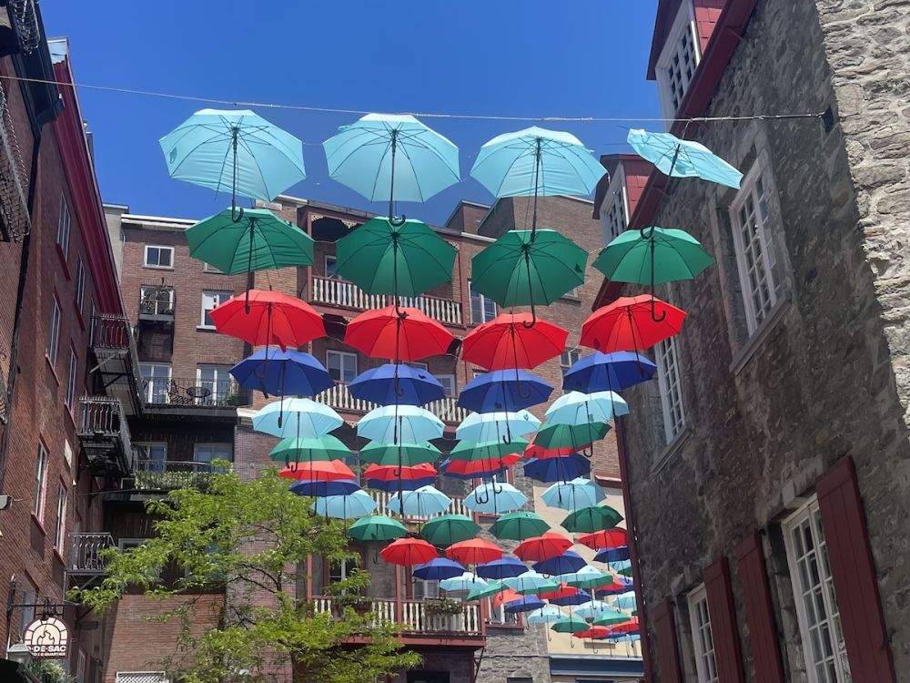 Rows of red, green, light blue, and dark blue umbrellas hang over a pedestrian alleyway between stone buildings