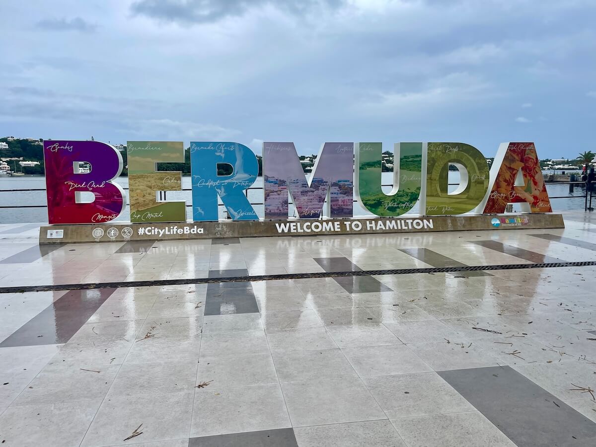 A large colorful sign that says 'BERMUDA,' and the words 'welcome to Hamilton' underneath. The sky is cloudy and the marble surface in front of the sky is slick with rain.