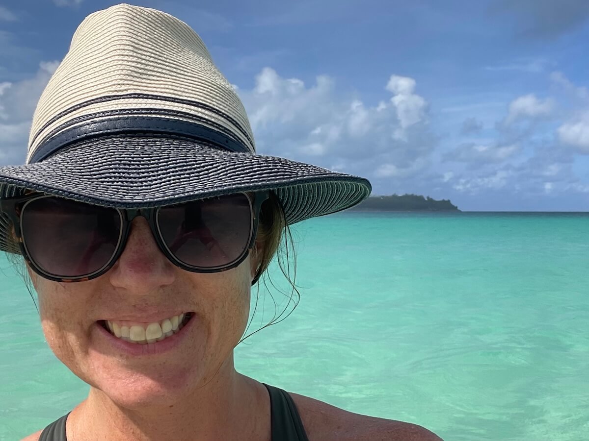 Lauren, a white woman in a blue wide-brimmed hat, sunglasses, and a green bathing suit, smiles at the camera. Incredibly clear blue-green water and a green spit of land can be seen behind her.