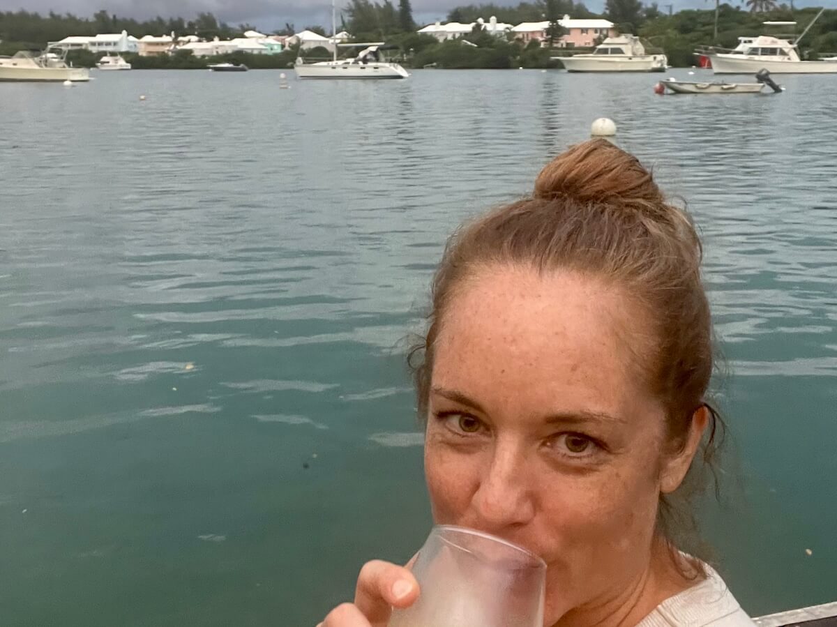 A selfie of Lauren, a white woman with brown hair pulled into a bun, smiling at the camera with a wine glass to her lips. She's sitting on a dock right at the water's edge, with boats and waterfront homes behind her.