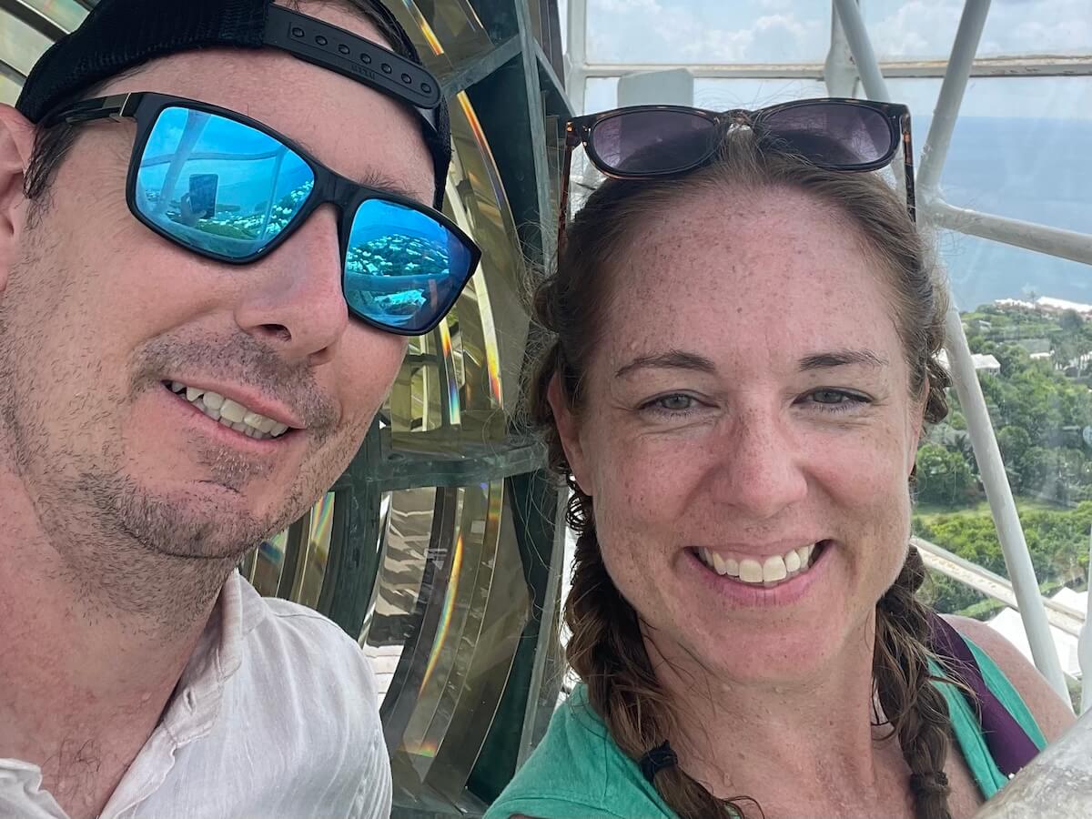 Jared, a white man, and Lauren, a white woman, smile at the camera as they stand next to a lantern at the top of a lighthouse. They are sweating, and the lush green and blue seas of Bermuda can be seen below through the window.