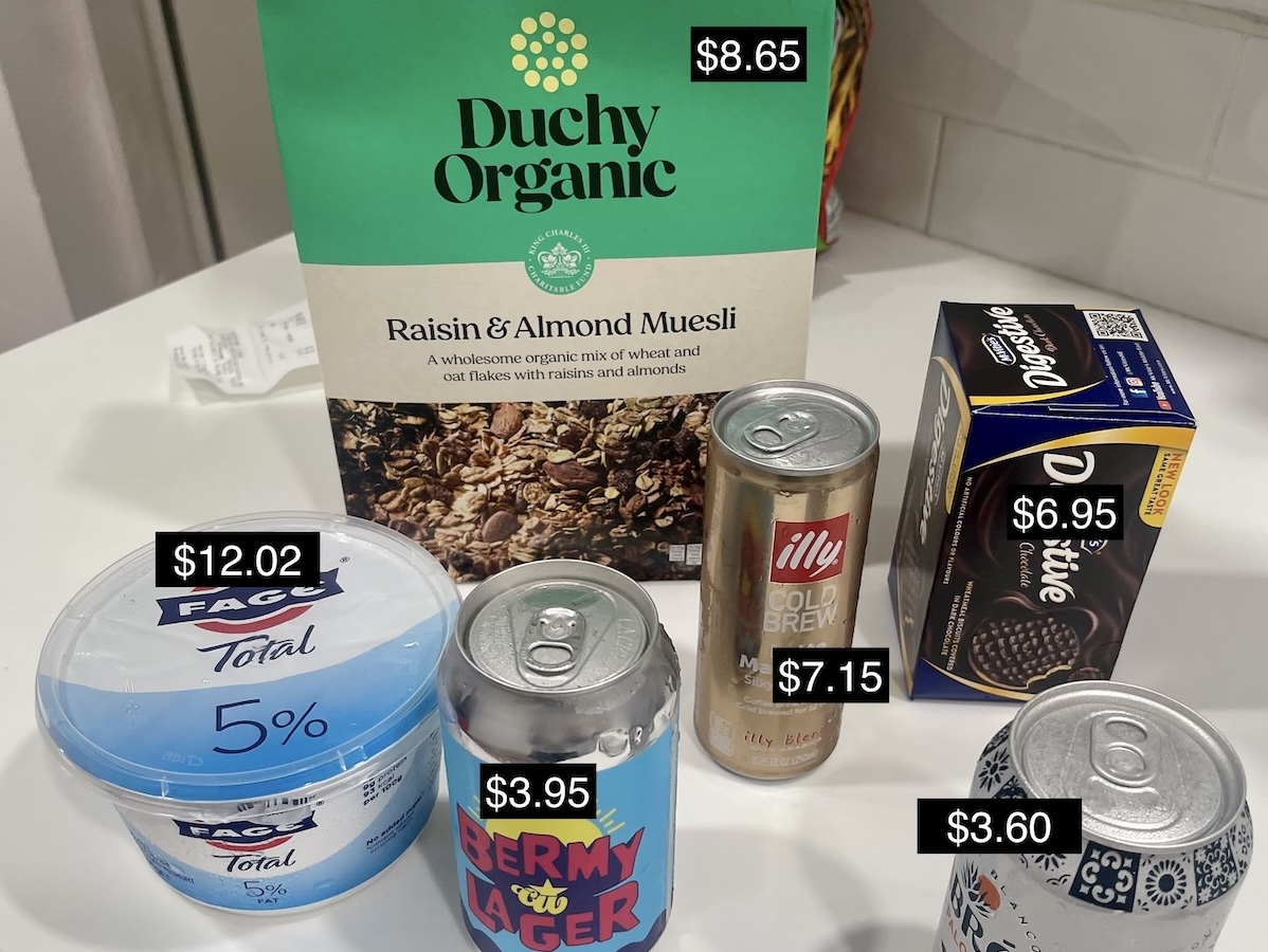 A photo of grocery items labeled with their prices: $8.65 for muesli, $7.15 for illy cold brew, $6.95 for digestive biscuits, $3.60 for a canned Paloma, $3.95 for Bermy Lager, and $12.02 for a small container of Greek yogurt.