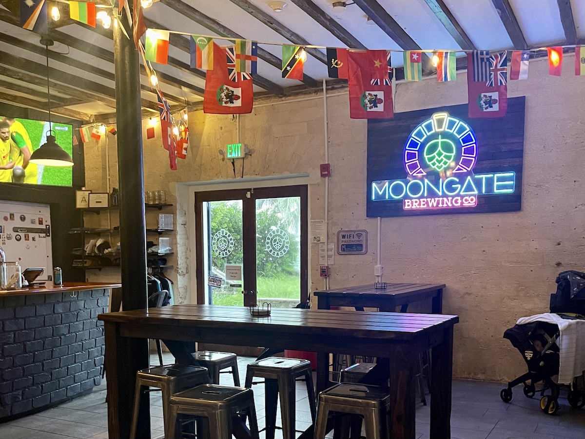 The interior of Moongate Brewing Co in Bermuda, a dark room with a neon logo sign, long wooden tables and stools, and a baby stroller in the corner.