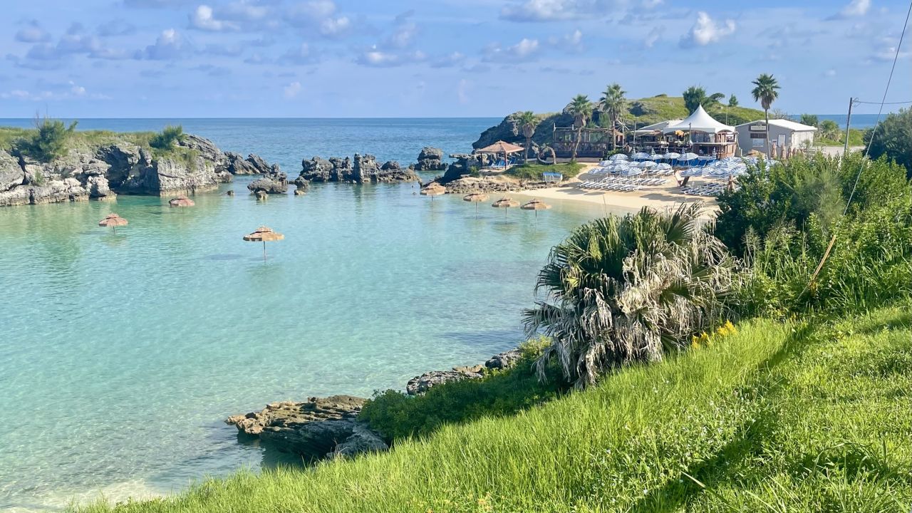 Clear blue bay curving into green grass, a small white beach, and a rocky border leading to the ocean.